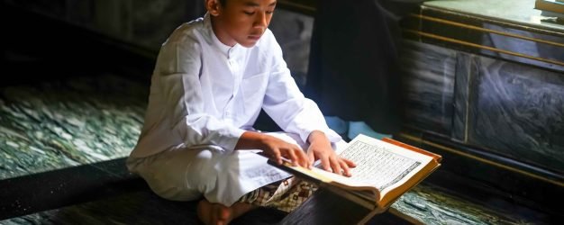 In a mosque, a young boy sits on the floor, focused intently on reading a religious text. Soft light illuminates the space, creating a serene atmosphere as others sit nearby in contemplation.